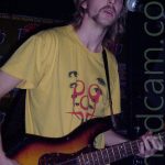 A man with long blond hair and a wispy beard, wearing a yellow t-shirt with black eyes in a v-shape overlaid by red swirls, playing a 4-stringed bass guitar. He is in a dark room with black walls, and standing beside a microphone on a stand. There is a green poster on the wall behind him advertising a New Years Eve party.