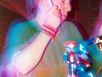 A man with ear-length brown hair and wearing a green polo-shirt and a blue pair of jeans, sitting behind a drum kit with a drum stick raised in the air in his right hand, looking down to his left. A striped sweat-band is wrapped around his right wrist. A green and chrome drumkit is sitting in front of him, with another drum and cymbal peeking in to the bottom left corner. A camera flash has frozen him in place, while a long exposure, his movements, and the stage lights paint red and green lights around him.