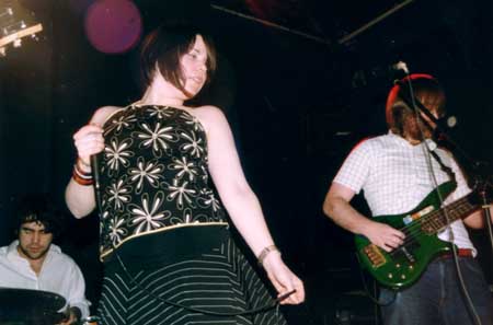 Low-down shot of a woman with brown hair cut in a bob and wearing a black and white flower-print top and black and white stripey skirt, holding a microphone in her right hand and looking off to her left. She is slightly off-centre, on the left of the image. Behind her and to her right is a man with long, curly dark hair and wearing an open-necked white shirt, sitting behind a drum kit. On the right of the frame is a man with dark, chin-length brown hair and a brown beard, wearing a checked white shirt and black jeans, playing a green bass guitar. The walls and ceiling behind them are almost entirely black, with a bright light shining in the top left corner.