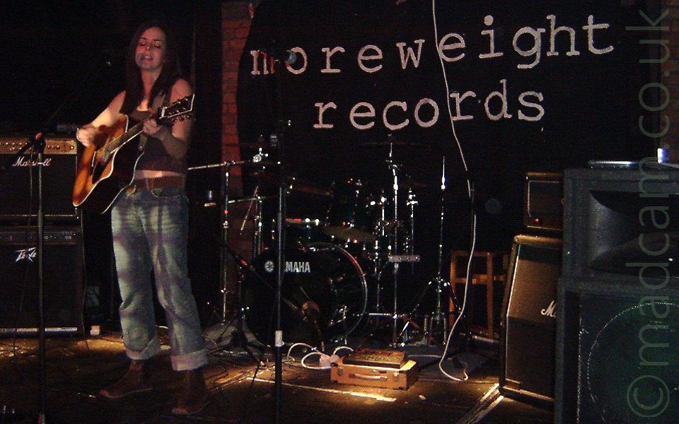 A woman with long, dark hair cascading around her shoulders, wearing a brown tank-top that slightly exposes her midriff, playing a brown acoustic guitar and singing into a microphone in front of her. She is standing off to the left of the photo, the rest of the image is taken up by a black and chrome drum kit in front of a wall with a black banner with white text "MoreWeight Records" hanging on it.