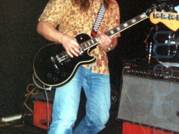 A man with long brown hair flowing over his shoulders, and a Cavalier-style moustache and goatee beard, wearing a brown flower-print shirt and a blue pair of jeans with black and grey trainers, playing a black electric guitar. There are grey and black amplifiers on upturned red crates behind him, with black walls and ceiling beyond that.