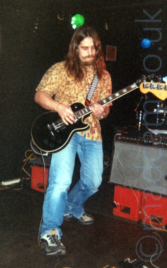 A man with long brown hair flowing over his shoulders, and a Cavalier-style moustache and goatee beard, wearing a brown flower-print shirt and a blue pair of jeans with black and grey trainers, playing a black electric guitar. There are grey and black amplifiers on upturned red crates behind him, with black walls and ceiling beyond that.