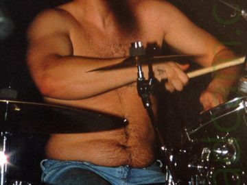 A shirtless man with dark hair and wearing a blue pair of jeans, sitting behind a black and chrome drumkit. Hisa arms are slightly raised as he plays his instrument, slightly leaning back his head turned slightly to his left with his eyes closed. Black walls fill the background.