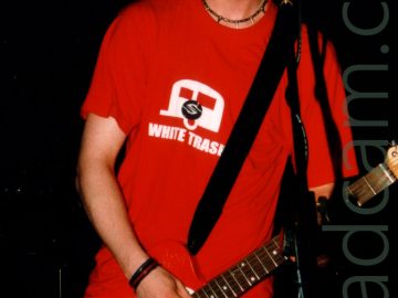 A man with short dark hair, wearing a blue baseball cap, a red t-shirt with a white caravan logo and the words "White trash", and a blue pair of jeans. A black strap slung over his left shoulder supports a red and white guitar he is playing with his left hand. He is standing behind a microphone mounted on a vlack stand. The space behind him is almost entirely black.