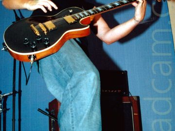 Low angle shot of a man with curly black hair and goatee beard, wearing a black tank-top and a blue pair of jeans with black boots, playing a black and amber wood electric guitar. A brown and black amplifier is on the floor behind him. He is casting a big shadow on the blue wall and white ceiling.