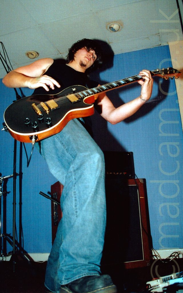 Low angle shot of a man with curly black hair and goatee beard, wearing a black tank-top and a blue pair of jeans with black boots, playing a black and amber wood electric guitar. A brown and black amplifier is on the floor behind him. He is casting a big shadow on the blue wall and white ceiling.