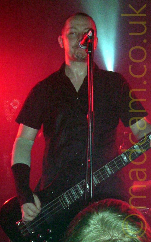 A man with close-cropped dark hair, wearing a black shirt and with his elbows and wrists strapped up, playing a black bass guitar and standing, grimacing, behind a microphone on a stand. Red light floods the background, with a single white light shining upwards behind his left shoulder. The head of a fan watching the gig is right at the bottom of the frame.