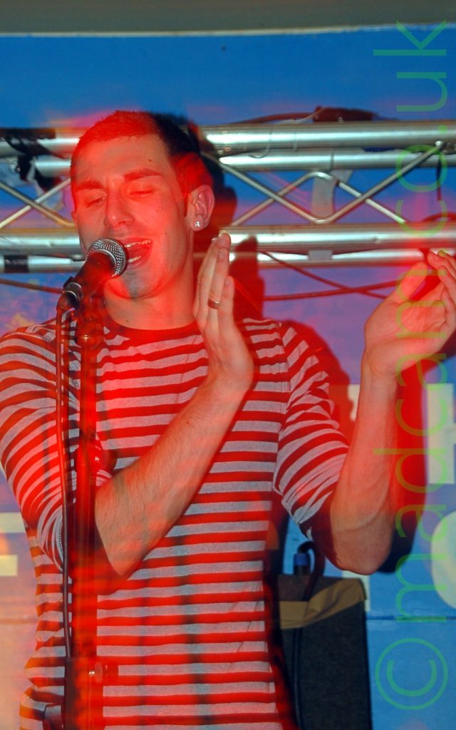 A man with dark, close-cropped hair and wearing a long-sleeved t-shirt with black and white bands around the body, with the sleeves rolled up to his elbows. He is singing into a microphone which is mounted on a stand in front of him, with his hands raised to hes left side as if clapping. The stage lights are casting a red aura around him, while the walls behind him are blue. A tubular metal framework can be seen stretching across the frame in the background.