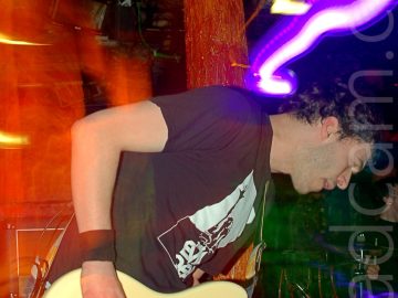 A man with short black hair, wearing a black t-shirt bearing a large, square, white and black logo, and a pair of blue jeans, playing a white, electric bass guitar. He is bent over, leaning to his left. A flash from the camera has frozen him in place, while a long exposure and a snap zoom has left an orange aura on the left of the frame, and streaks of yellow and purple light on the right. An orange pole with several faces on it, painted and glazed to look almost organic, stands behind him, while the bands drummer can be seen in the bottom right of the frame. Dark walls and ceiling fill the rest of the frame.