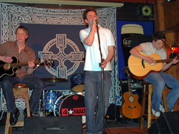 3 men performing on a stage as part of a band. On the left is a man with short blond hair, wearing a brown shirt and a pair of black jeans, sitting on a tall brown stool and playing a black acoustic guitar. In the middle is a man with spiky brown hair and weraing a white polo short and grey jeans, singing into a microphone on a stand, holding the mic in his right hand and the stand in his left. On the right is a man with short dark hair, wearing a white t-shirt and a blue pair of jeans, sitting on a tall wooden stool and playing a brown acoustic guitar. In the background in the middle is a sparkly blue and chrome drum kit, with a large, black "4*Mary" logo in the middle of the bass drum skin, with another brown acoustic guitar off to one side. On the wall is a large, dark blue fabric covering with a large, white Celtic cross in the middles, and bordered with elaborate white scrollwork. Large blue posters featuring over-sized pint glasses filled with a black liquid with a white top, and emblazoned with the word "Guiness" are on either side of the stage.