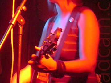 A woman with dark, chin-length hair cut in a shaggy bob, wearing a brown tank-top and denim skirt, playing an acoustic guitar, and singing into a microphone on a stand in front of her. A long exposure has been used, her right hand a blur as she strums her guitar, while the stage light cast an orange hue over everything. Dark walls and ceiling fill the background.