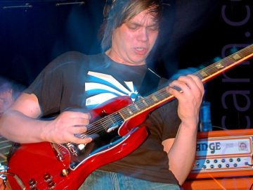 A man with mid-length dark hair, wearing a black t-shirt with a square, black and white sunburst logo in the middle, and a blue pair of jeans, playing a red and black electric guitar. He is leaning back slightly, looking down at his guitar with a slightly aggressive look on his face. A camera flash has frozen him in place, with a long exposure creating movement, as well as blue or purple secondary images creating haloes of light. An orange and black amplifier is behind him in the right of the frame, with a red and chrome drum kit on the left, with black walls and ceiling beyond that.