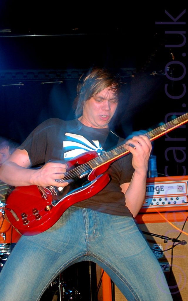A man with mid-length dark hair, wearing a black t-shirt with a square, black and white sunburst logo in the middle, and a blue pair of jeans, playing a red and black electric guitar. He is leaning back slightly, looking down at his guitar with a slightly aggressive look on his face. A camera flash has frozen him in place, with a long exposure creating movement, as well as blue or purple secondary images creating haloes of light. An orange and black amplifier is behind him in the right of the frame, with a red and chrome drum kit on the left, with black walls and ceiling beyond that.