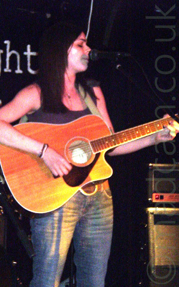 A woman standing on a stage, with long dark hair draped around her shoulders, with a brown vest top and a blue pair of jeans, playing a brown acoustic guitar and singing into a microphone on a stand in front of her. Black walls fill the background.