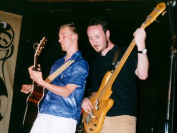 2 members of a band performing on stage. On the left is a man with short blond hair, wearing a blue, short sleeved shirt with white flecks, and white shorts, playing a brown acoustic guitar, with a tan guitar strap slung over his left shoulder. On the right is a man with short dark hair and a straggly beard, wearing a black polo shirt and light brown trousers, playing a pale-coloured bass guitar. Tan sheets of cloth with ornate black decorations cover sections of the black walls in the background.