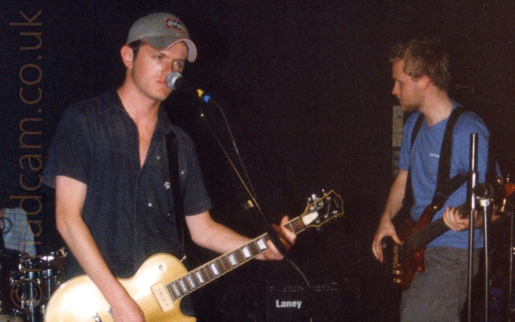 2 members of a band performing on a stage. On the left is a man with short dark hair under a grey baseball cap, wearing a dark blue shirt, and playing a gold-coloured electric guitar. He is glowering at the microphone on a stand in front of him. On the right of the frame is a man with short brown hair and a thin beard, wearing a blue t-shirt and grey grousers, with a brown bass guitar attached to a black shoulder harness. Black walls fill the rest of the frame.