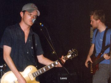 2 members of a band performing on a stage. On the left is a man with short dark hair under a grey baseball cap, wearing a dark blue shirt, and playing a gold-coloured electric guitar. He is glowering at the microphone on a stand in front of him. On the right of the frame is a man with short brown hair and a thin beard, wearing a blue t-shirt and grey grousers, with a brown bass guitar attached to a black shoulder harness. Black walls fill the rest of the frame.