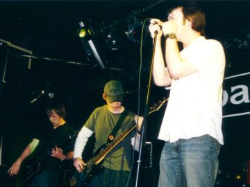 3 members of a band performing on a stage. On the left is a man with mid-length hair, wearing a black shirt and dark blue jeans, playing a black electric guitar, looking down towards his instrument. In the middle is a man wearing a khaki green hat, a green t-shirt over a white jumper, and a blue pair of jeans, playing a black bass guitar. On the right is a man with short brown hair, wearing a white shirt and a blue pair of jeans, holding on to a mic stand as if his life depends on it. Black walls and ceiling fill the rest of the frame, with large white letters "AR" on the rear wall, part of the word "Barfly" on the wall on the right of the frame.