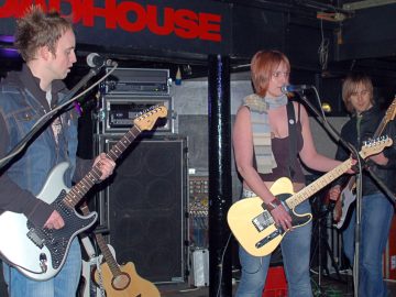 3 members of a band on a stage. On the left is a man with short, brown, spiky hair, wearing a sleeveless denim jacket over a black hoodie and blue jeans, and playing a grey and black electric guitar. In the middle is a woman with her hair cut in a brassy bob, wearing a grey woollen scarf over a strappy brown top that reveals her midriff and a pair of blue jeans, playing a yellow-ish electric guitar and singing in to a microphone on a stand in front of her. On the right is a man with shaggy brown hair, wearing a black leather jacket over a green t-shirt and blue jeans, playing a brown and white bass guitar. Racks of amplifiers line the black walls behind them, with a brown acoustic guitar casually leaning against one of the amps.