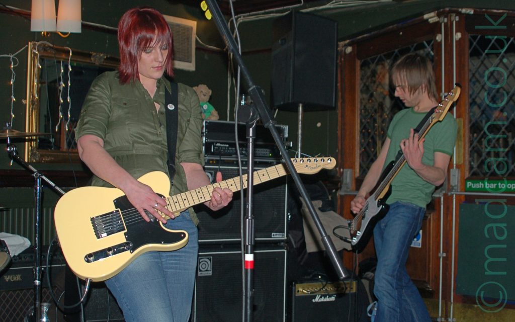 2 members of a band performing on stage. On the left is a woman with shoulder-length red hair, wearing a green shirt and a blue pair of jeans, playing a cream-coloured electric guitar. A tall microphone stand is in front of her. On the right is a man with brown shaggy hair, wearing a green t-shirt and blue jeans, playing a black and white bass guitar, facing off to the left of the frame. A collection of black amplifiers litters the stage behind them, with 2 glazed, brown wooden doors on the right of the frame. A heavily gilded mirror hangs on the green wall under a pair of white lampshades on the left of the frame. Green walls and ceiling fill the rest of the frame.
