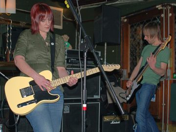 2 members of a band performing on stage. On the left is a woman with shoulder-length red hair, wearing a green shirt and a blue pair of jeans, playing a cream-coloured electric guitar. A tall microphone stand is in front of her. On the right is a man with brown shaggy hair, wearing a green t-shirt and blue jeans, playing a black and white bass guitar, facing off to the left of the frame. A collection of black amplifiers litters the stage behind them, with 2 glazed, brown wooden doors on the right of the frame. A heavily gilded mirror hangs on the green wall under a pair of white lampshades on the left of the frame. Green walls and ceiling fill the rest of the frame.