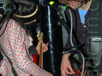 Two members of a rock band performing on a stage. On the left is a woman with long dark hair with a woven yellow hair band, wearing a white dress with a red and black floral pattern, and playing a red electric guitar. On the right is a man with mid-length shaggy hair, wearing a black shirt over a blue t-shirt, and dark coloured trousers, playing a black electric bass guitar. They are standing facing each other, and their heads are slightly bowed. A pair of microphone stands are in front of them in the foreground, while a glossy black metal pole holds up the ceiling immediately behind them. Amplifiers line black walls in the background.