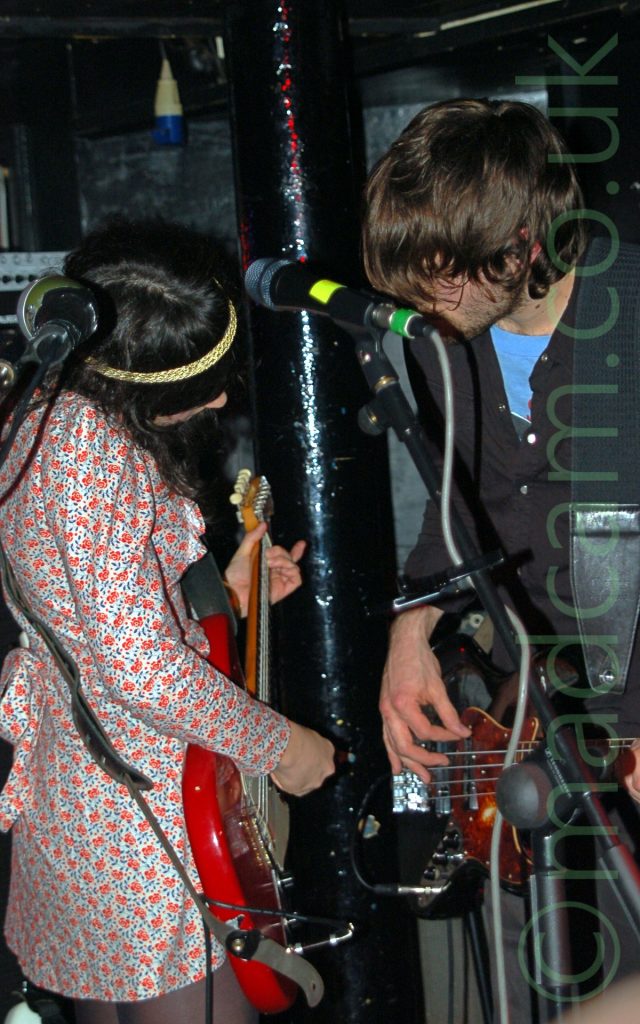 Two members of a rock band performing on a stage. On the left is a woman with long dark hair with a woven yellow hair band, wearing a white dress with a red and black floral pattern, and playing a red electric guitar. On the right is a man with mid-length shaggy hair, wearing a black shirt over a blue t-shirt, and dark coloured trousers, playing a black electric bass guitar. They are standing facing each other, and their heads are slightly bowed. A pair of microphone stands are in front of them in the foreground, while a glossy black metal pole holds up the ceiling immediately behind them. Amplifiers line black walls in the background.