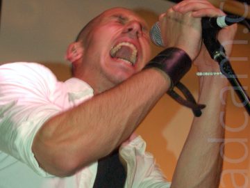 A man with a shaved head, wearing a white shirt with only the bottom few buttons done up over a black t-shirt, and with a leather wrist-band on his right wrist, leaning into frame from the left. He is holding on to a microphone on a stand with both hands, and is screaming in to the mic, his mouth wide open and showing lots of teeth, mostly on his upper jaw. He is casting a strong shadow on the cream coloured wall behind him.