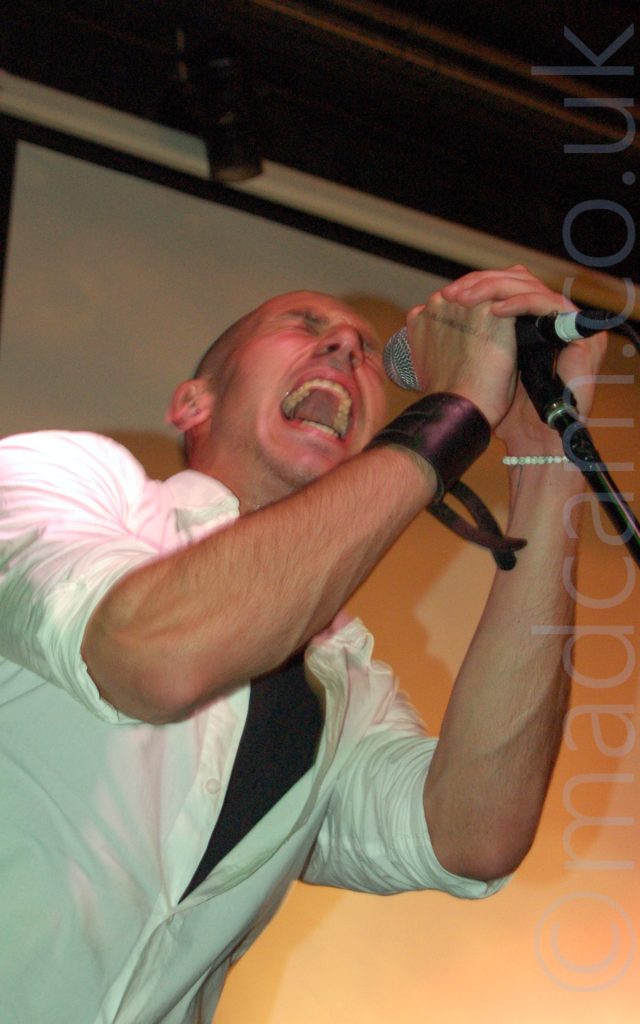 A man with a shaved head, wearing a white shirt with only the bottom few buttons done up over a black t-shirt, and with a leather wrist-band on his right wrist, leaning into frame from the left. He is holding on to a microphone on a stand with both hands, and is screaming in to the mic, his mouth wide open and showing lots of teeth, mostly on his upper jaw. He is casting a strong shadow on the cream coloured wall behind him.