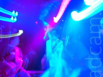 Close-up shot of a man with mid-length dark hair and wearing a light-coloured shirt, playing a bass guitar. A long exposure and a snap zoom means he appears twice in the photos, bathed in blue light, the second time appearing far away. On the left of the frame, another member of the band is playing an electric guitar, illuminated by a purple light. Streaks of blue and purple light flood the top half of the image.