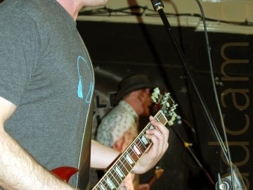 Side view of a man with shaggy brown hair, wearing a grey t-shirt with a blue headphones logo in the middle, facing to the the right. He is playing a red electric guitar, and singing in to a microphone on a stand in front of him. In the background is a man wearing a black, wide-brimmer hat, a white shirt with a blue floral pattern, and a pair of grey trousers. He is playing a grey electric guitar, and singing into a microphone on a stand. Black walls and white ceiling fill the rest of the frame.