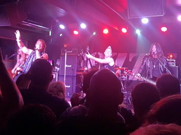 5 members of a band on stage. On the left is a man with long hair, wearing a black t-shirt, and playing an electric guitar. To his right and slightly obscuring him is a man with shoulder-length hair, wearing a sleeveless jacket and blue scarf, with his right arm raised in the air and a microphone held in his left hand, which he is singing in to. In the centre of the image is a man with short blond hair teased up into a single spike, wearing a black sleeveless t-shirt, and playing a electric guitar which is being swung around with reckless abandon. Behind him, almost hidden away, is a man wearing a sleeveless black t-shirt, sitting behind a drum kit. On the right is a man with long, dark hair, wearing a leather jacket over a black t-shirt, and with a large cross on a chain around his neck, playing an inverted bass guitar in a left-handed position. He is singing into a microphone on a stand in front of him. The heads of people in the crowd fill the foreground, with a black backdrop with the white text "Fozzy" stretched between 2 stacks of amplifiers in the background, with bright purple, red, and orange stage lights attached to the black roof above them. The whole scene has a purple cast from these lights.