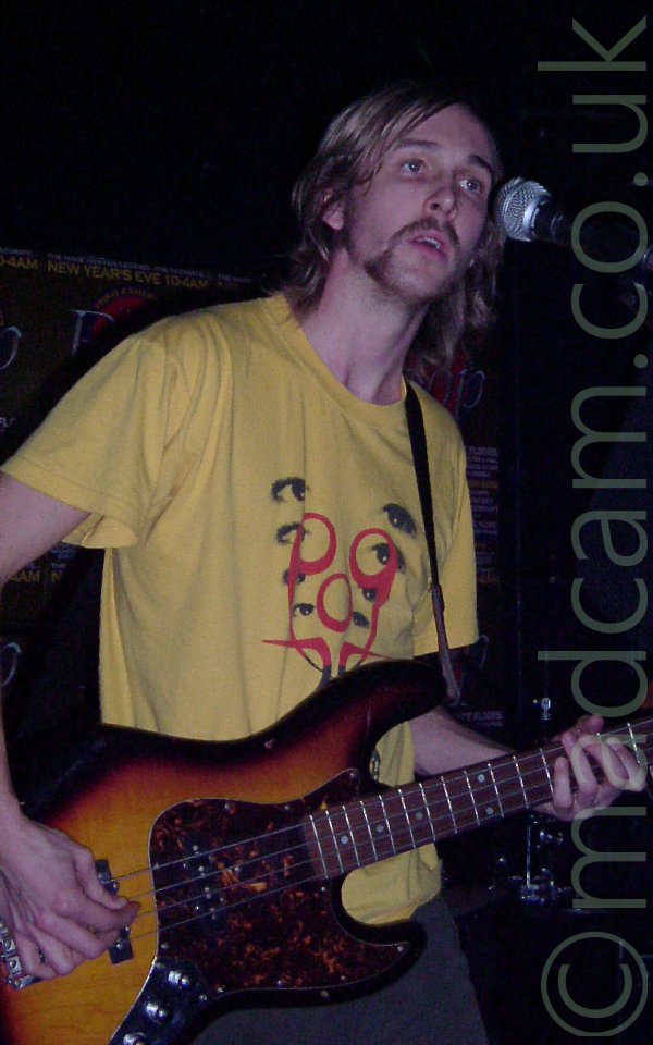 A man with long blond hair and a wispy beard, wearing a yellow t-shirt with black eyes in a v-shape overlaid by red swirls, playing a 4-stringed bass guitar. He is in a dark room with black walls, and standing beside a microphone on a stand. There is a green poster on the wall behind him advertising a New Years Eve party.