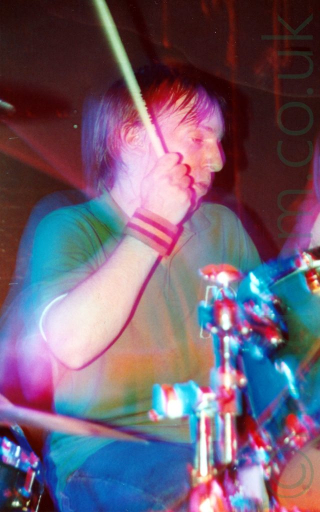 A man with ear-length brown hair and wearing a green polo-shirt and a blue pair of jeans, sitting behind a drum kit with a drum stick raised in the air in his right hand, looking down to his left. A striped sweat-band is wrapped around his right wrist. A green and chrome drumkit is sitting in front of him, with another drum and cymbal peeking in to the bottom left corner. A camera flash has frozen him in place, while a long exposure, his movements, and the stage lights paint red and green lights around him.