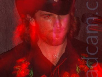 A man with shoulder-length brown hair, wearing a black stetson hat, and a black shirt with red piping, and red flowers across the chest. A flash has frozen him in place, while a long exposure and movement have combined with stage lights to paint red light around his head. A black wall behind him fills the rest of the frame.