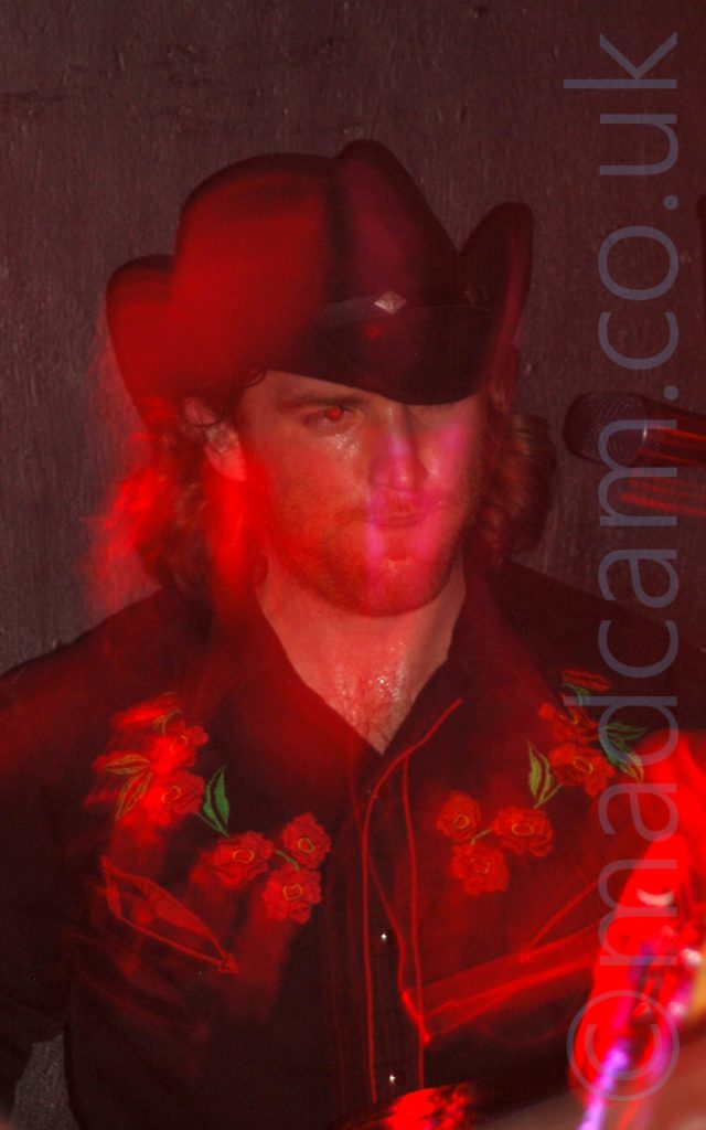 A man with shoulder-length brown hair, wearing a black stetson hat, and a black shirt with red piping, and red flowers across the chest. A flash has frozen him in place, while a long exposure and movement have combined with stage lights to paint red light around his head. A black wall behind him fills the rest of the frame.