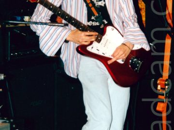 A man with long, wavy black hair, wearing an open white shirt with red and blue pin-stripes and large, open cuffs on the sleeves, over a black t-shirt with a white logo, and flared white jeans, playing a red and white guitar left-handed. He is looking down at his guitar, his face covered with his long hair. A microphone on a stand is in front of him, but he isn't using it. There are a number of black amps scattered around behind him, leaning against the orange and yellow wall, or the black wall at the back of the stage