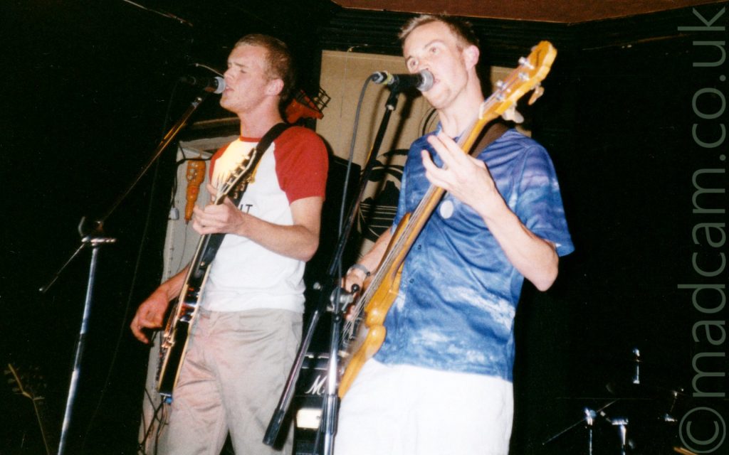 Two men in a band, playing electric guitars and singing in to microphones on stands in front of their faces. On the left is a tall man with short blond hair and wearing a white t-shirt with red shoulders and sleeves and pale trousers, and playing a black guitar with his right hand, holding it in his left as he sings into a mic. On the right is a man with short, slightly spiky blonde hair, wearing a blue t-shirt and white pants, playing a yellowy-brown bass guitar held in his left hand, and singing into a mic. The walls of the room behind them are black, with a sandy brown backdrop on one wall, and a red ceiling.
