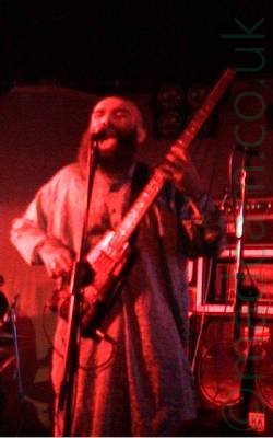A man with a bald head and big, bushy, black beard, wearing Shalwar Kameez (a traditional form of dress worn in East Asia, particularly Pakistan, Afghanistan, and parts of India), and playing a black headless bass guitar. He is singing into a microphone on a stand in front of him. In the background, racks of amplifiers can be seen, with curtains covering the walls behind them. The whole image has a red tint from the stage lights.