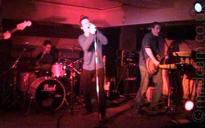 3 members of a band performing on a stage, on the left is the drummer at the back of the stage, with short dark hair and a grey and black t-shirt. In the middle is a man with short dark hair, wearing a grey jumper and black trousers, leaning on a microphone stand as he sings into the mic. On the right is a man with short dark hair, wearing a grey t-shirt and dark trousers, playing an electric guitar, and singing into a microphone on a stand, behind a bank of keyboards. The neck of a bass guitar can be seen right on the left of the frame, with a hand holding it half way along. The room is lit by a group of red lights, with shadows reaching out in all directions.
