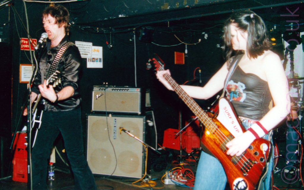 2 members of a band performing on a stage. On the left is a man with brown hair, wearing a satin shirt and dark blue trousers, playing a guitar and shouting in to a microphone on a stand in front of him. On the right is a woman with long dark hair and wearing a black top that rises over her right shoulder and blue jeans, playing a brown bass guitar with her left hand, looking closely at her right hand on the neck of the bass. In the background, amplifiers and an assortment of red crates stand against the black walls.
