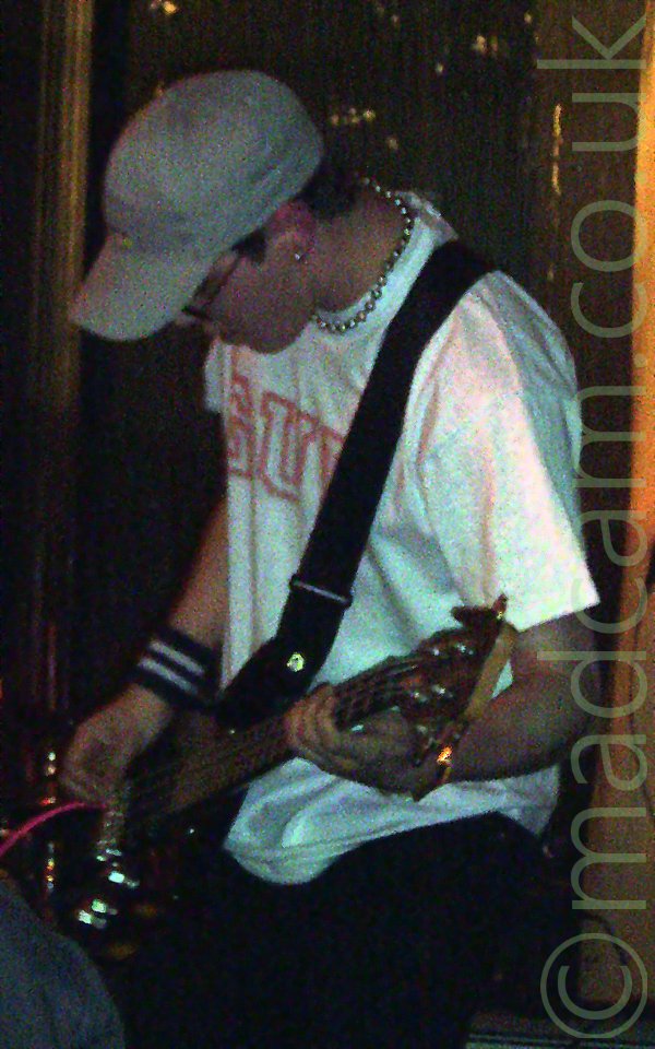 Photo of a man wearing a white t-shirt with some red text, and a pale blue cap, playing a bass guitar. He is facing to the left, and has a black and white band around his right arm, just above the wrist,, and a necklace around his neck. The walls behind him are reflecting golden light.