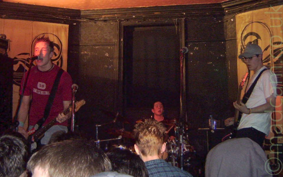 3 members of a rock band performing on a stage. On the left is a man with spiky brown hair, wearing a red t-shirt with a white logo across the chest, and playing an electric guitar whilst singing into a microphone on a stand in front of him. In the middle, right at the back of the stage, the brown-haiored head of a man floats above a drum kit. On the right, a man with a pale blue baseball cap and a pair of glasses, and wearing a white t-shirt and dark trousers is playing a bass guitar. In the foreground, the backs of the heads of some fans can be seen down in the pit. In the background, the centre section of wall is all black, with outer sections at a slight angle are covered with gold-coloured drapes with a large black logo.