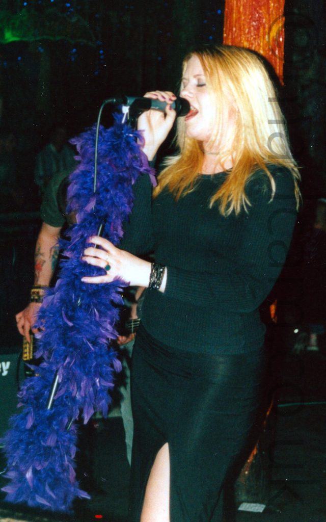 Photo of a woman with shoulder-length blonde hair and wearing a black top and black skirt with a long slit on the left leg. She is singing into a microphone on a stand, the stand wrapped in a purple feather boa, holding the mic in her right hand and the stand in her left, with her eyes closed. A wooden pole painted a bright orange and red is behind her head, and a tattooed arm holding a black bass guitar is just visible on the left of the frame.