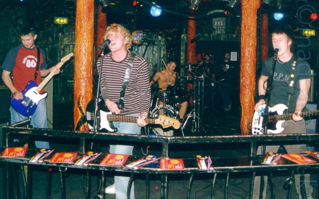 4 members of a band on stage. On the left is a man with short dark hair, wearing a red top with blue shoulders and sleeves and a white logo, playing a purple and white guitar. In the middle is a man with curly blond hair, wearing a white and brown banded jumper and pale blue jeans, with a green bass guitar slung over his left shoulder, scre ming into a microphone on a stand in front of him. Behind him is a man with short dark hair, with no top on and hopefully wearing shorts, playing a black and chrome drum kit, a stick held aloft in his left hand. On the right is another man with short dark hair, wearing a black t-shirt and dark grey trousers, playing a white and black guitar and singing into a mic on a stand in front of him. In the foreground, a black metal barrier stretches across the frame, a shelf on it strewn with red and black flyers. In the background, metal poles with moulded and painted surfaces glisten wetly, red and orange and yellow, resembling something organic and unspeakable. The dark, cave-like walls and ceiling add to the look. 2 men in the background wearing dark t-shirts and jeans are leaning against a poorly disguised door.