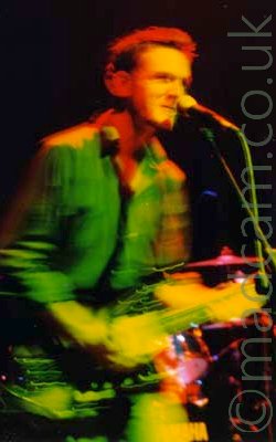 A man on stage, playing an electric guitar and singing into into a microphone in front of him, wearing an open-necked shirt. He is illuminated by various coloured lights - red, yellow, green, and his arms are blurred into smears as he moves during a long exposure. Cymbals and part of the drum kit can be seen behind him, illuminated in red, with the rest of the background being pure black.