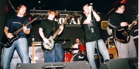 5 men standing on a stage, all members of a band. On the left is a man with long brown hair, wearing a blue t-shirt, blue jeans, and wielding a black guitar. Next to him is a man with long brown hair, wearing a black t-shirt and blue jeans, playing a black and grey bass guitar. At the back, a man with short dark hair and wearing a grey t-shirt is sitting behind a red and chrome drum kit. Next is a man with short brown hair, wearing a black t-shirt and pale blue trousers. He is holding a microphone mounted on a stand in both hands to his mouth, singing in to it. On the right is a man with short dark hair, wearing a black sweater and grey trousers, playing a black electric guitar. The walls and ceiling behind them are black, with a metal lighting rig running across the top of the back wall.