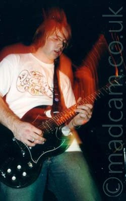 Long exposure shot of a man with dark hair, wearing a white t-shirt with a grey logo on the chest and blue jeans, playing a black guitar attached to a black strap slung over his left shoulder. A flash has frozen him in place, the long exposure capturing movement and stage lights afterwards, with a red secondary image of him leaning back slightly with his guitar raised, highlighted by red lights. Black walls and ceiling fill the rest of the frame.