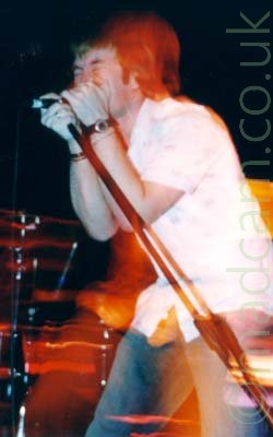 A man with dark hair and wearing a white shirt and blue pair of jeans, screaming into a microphone held on a stand in front of him. He is cupping the mic in both hands, leaning on the stand so it is slanted to the side. A flash has frozen him in place, while a long exposure shows movement after the shutter was triggered, with red and yellow secondary images around him. Black walls fill the background.