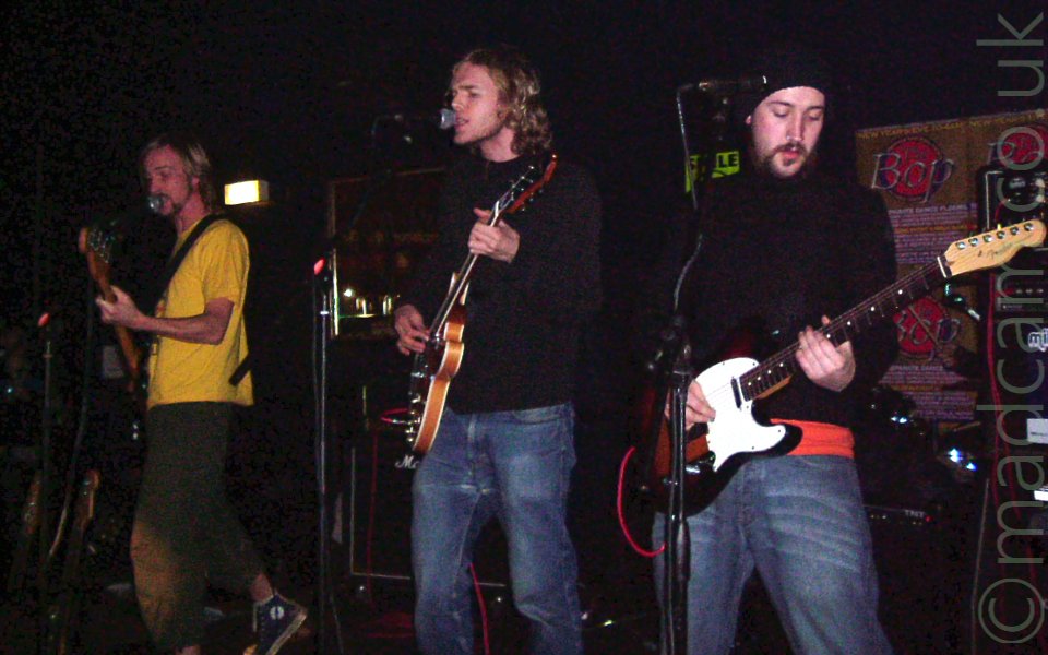 3 members of a band on stage. On the left is a man with long blond hair and beard, wearing a yellow t-shirt and dark trousers, playing a black bass guitar and singing in to a microphone on a stand in front of him. In the middle is a man with long blond hair, wearing a black jumper and blue jeans, playing a brown electric guitar and singing in to a mic in front of him. On the right is a man with a brown beard, a black woolly hat, black jumper over a red t-shirt, and blue jeans, playing a brown and white electric guitar. He isn't singing, although there is a mic on a stand in front of him, but instead is looking down at his guitar. There are rows of amplifiers behind them, with posters on black walls.